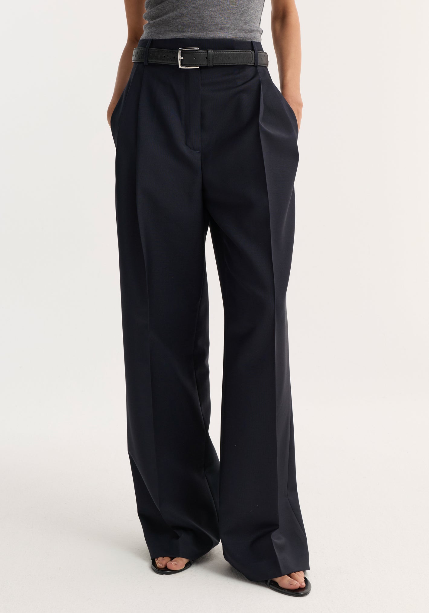 Person modeling mid-waist tailored trousers with wide-leg, front pleats, and side pockets, styled with a belt and heeled sandals against a white background.