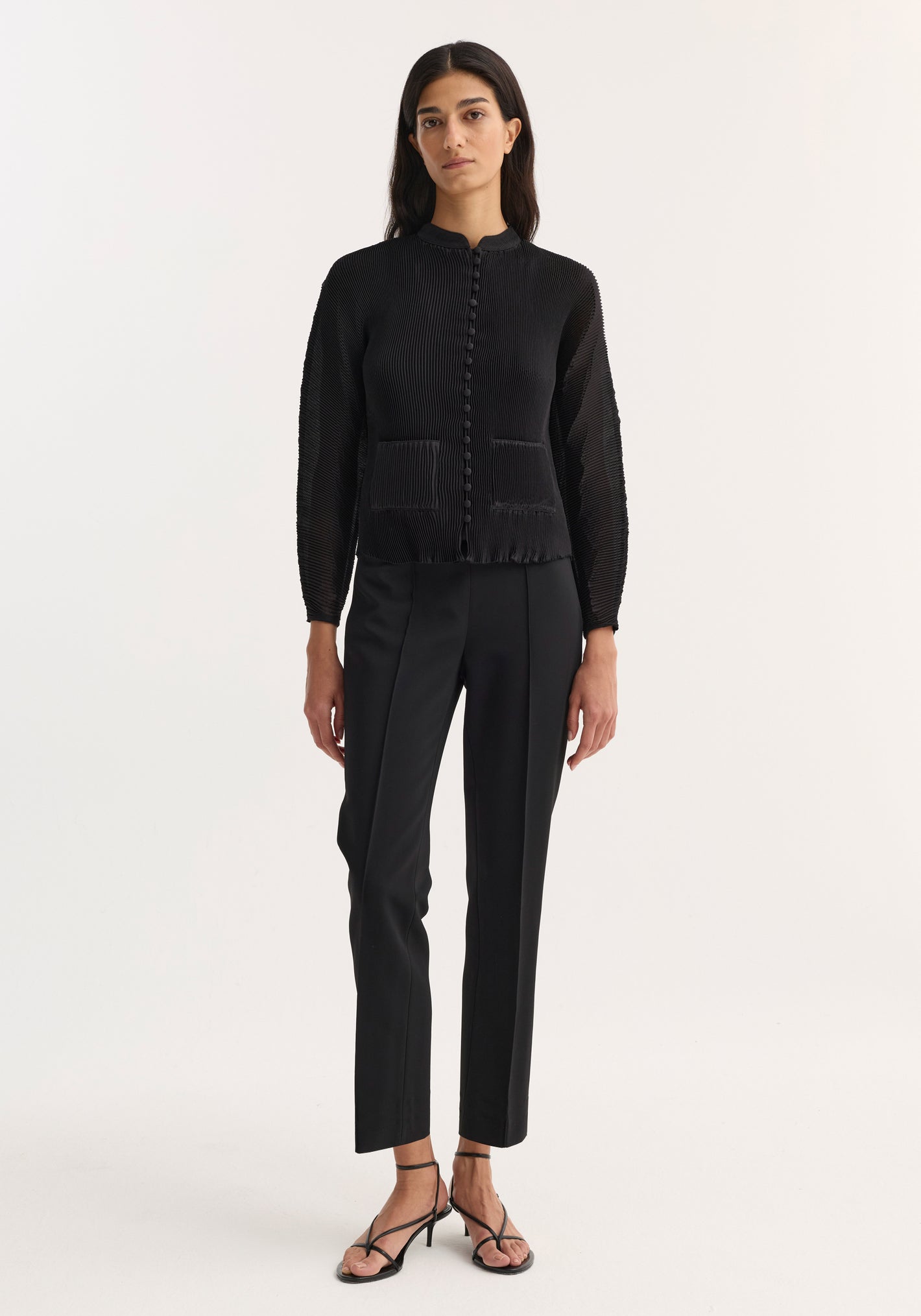 Model wearing Plissé cardigan in black, featuring a stand-up collar, button front, patch pockets, paired with tailored trousers and strappy sandals.