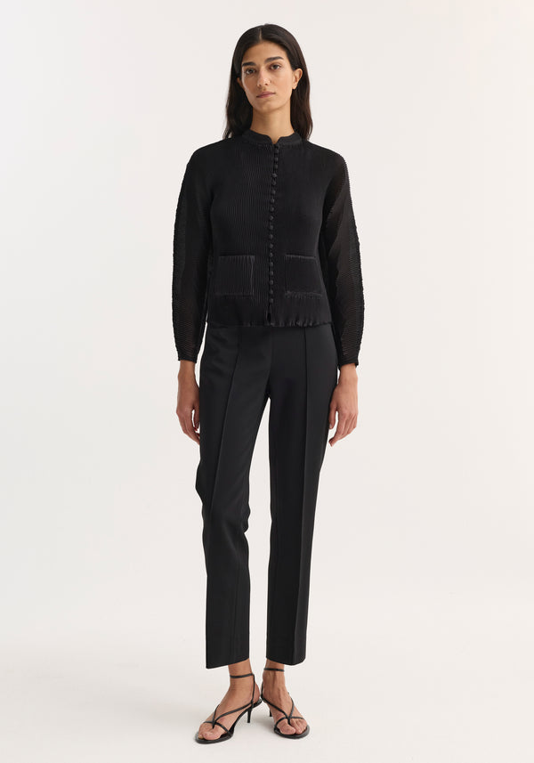 Model wearing Plissé cardigan in black, featuring a stand-up collar, button front, patch pockets, paired with tailored trousers and strappy sandals.