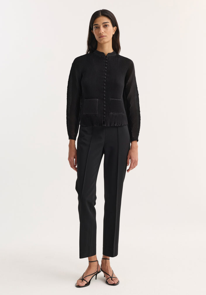 Model wears Cigarette leg trousers in black, featuring a straight narrow leg and high-rise fit, paired with a pleated jacket and strappy sandals.