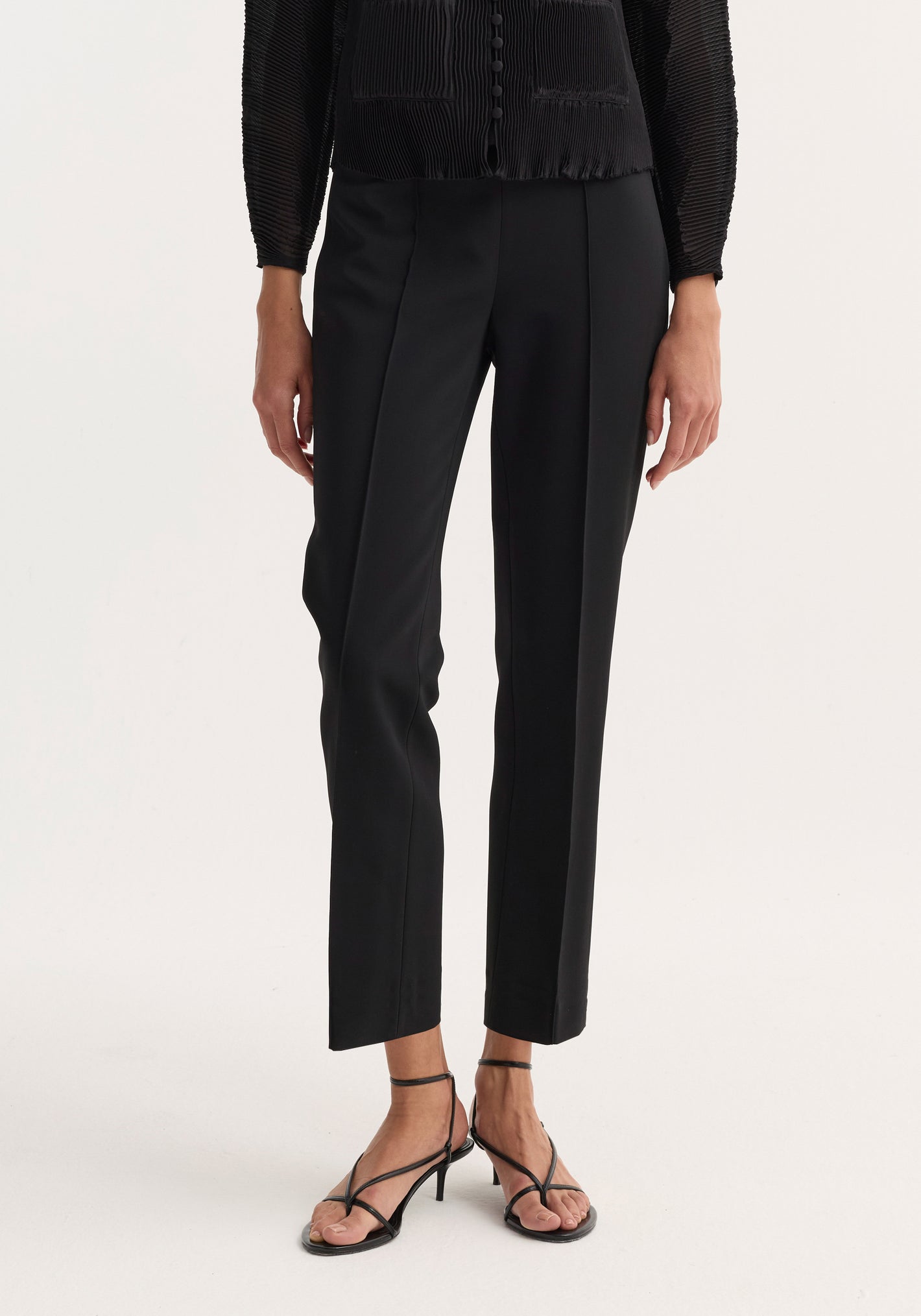 Cigarette leg trousers in black, featuring a pin-tuck crease and ankle length. High-rise fit, paired with strappy sandals, showcasing a sleek, versatile style.
