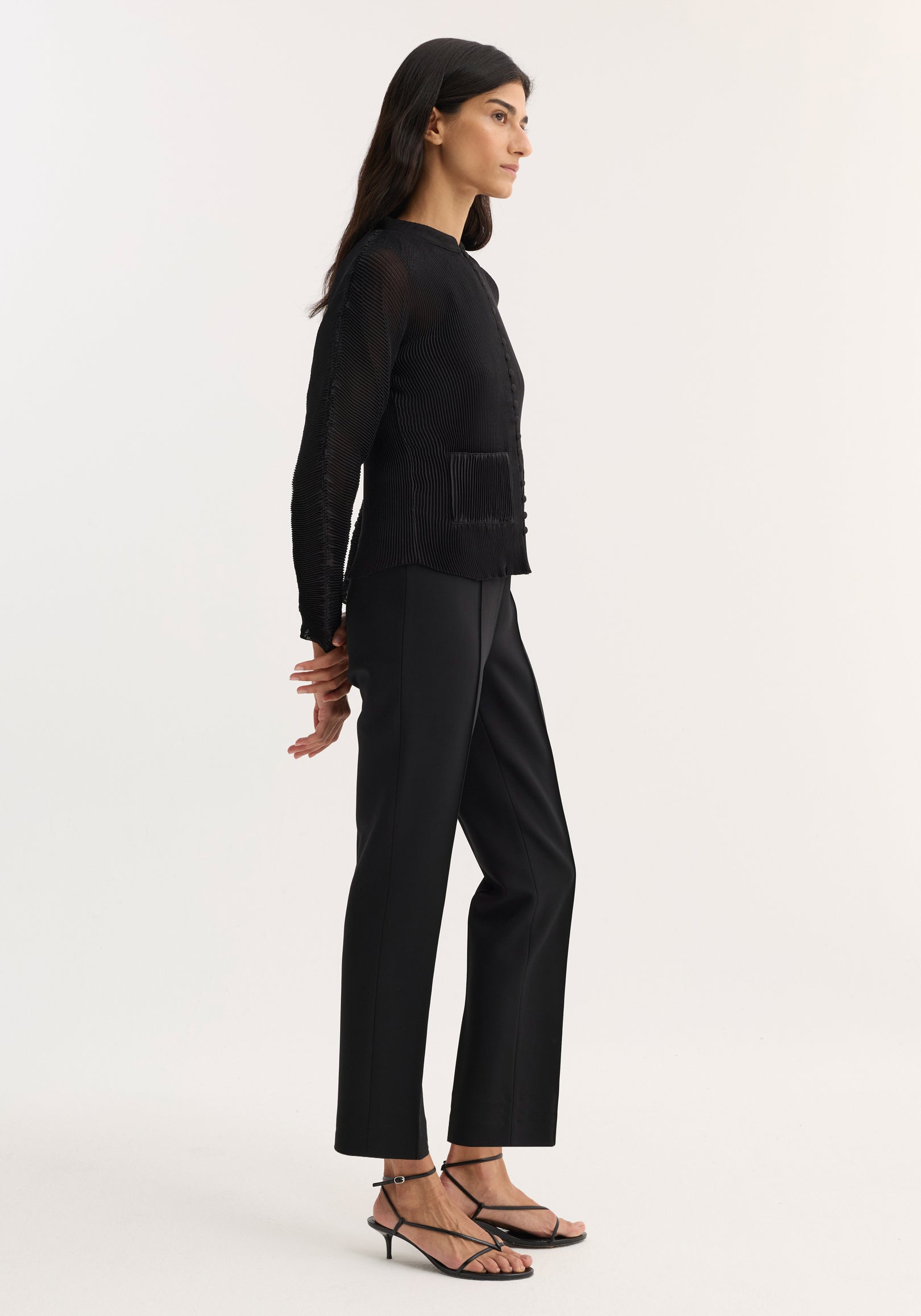 Woman wearing a black Plissé cardigan | black with long sleeves and front pockets, paired with black tailored pants and strappy black sandals, standing in profile against a plain background.