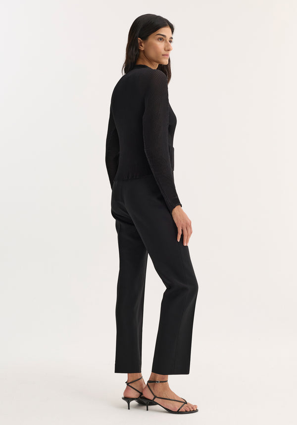 Woman wears Plissé cardigan, featuring a structured fit, stand-up collar, and patch pockets, paired with straight-leg trousers and low-heeled sandals.
