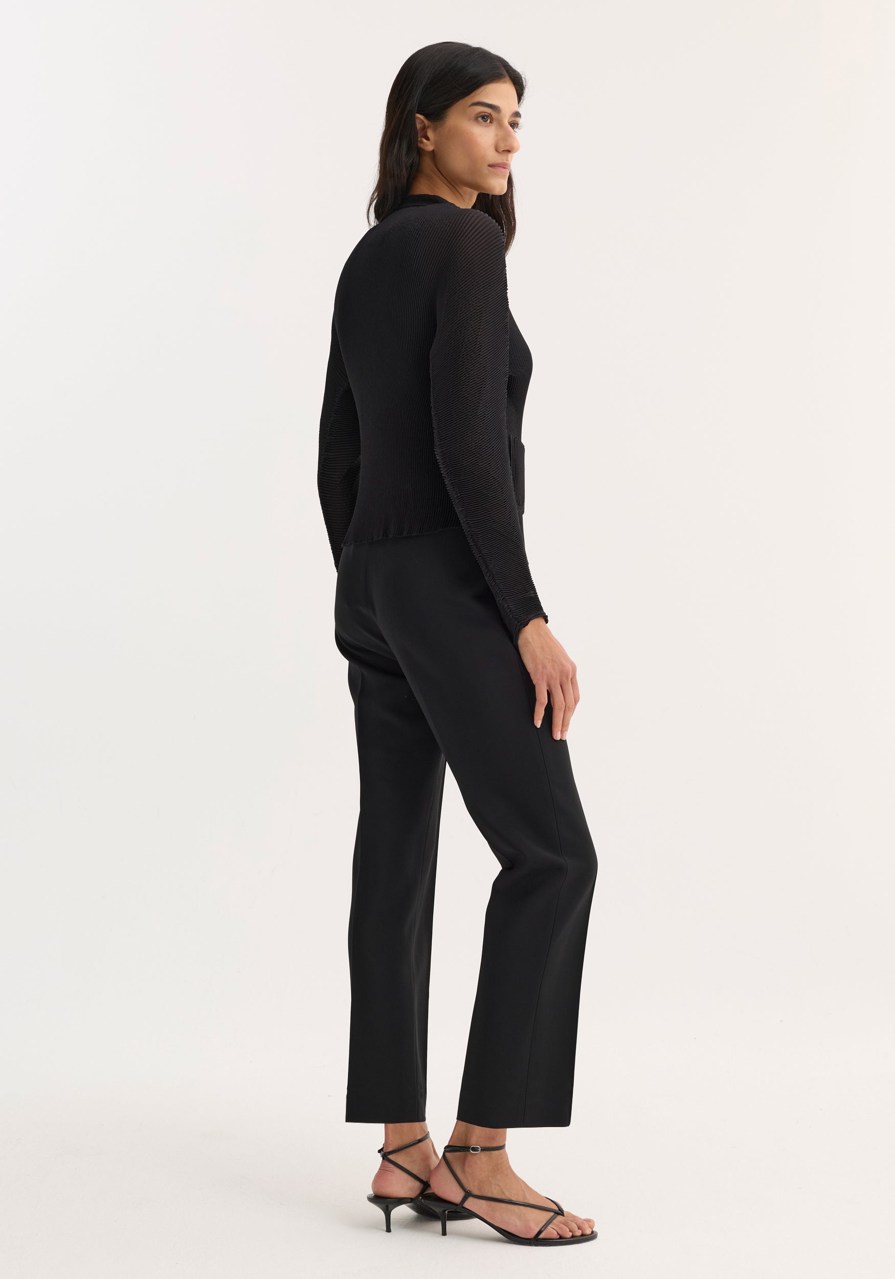 Woman wearing a black Plissé cardigan | black with long sleeves, paired with black pants and strappy black heels, standing in a side-back pose against a plain background.