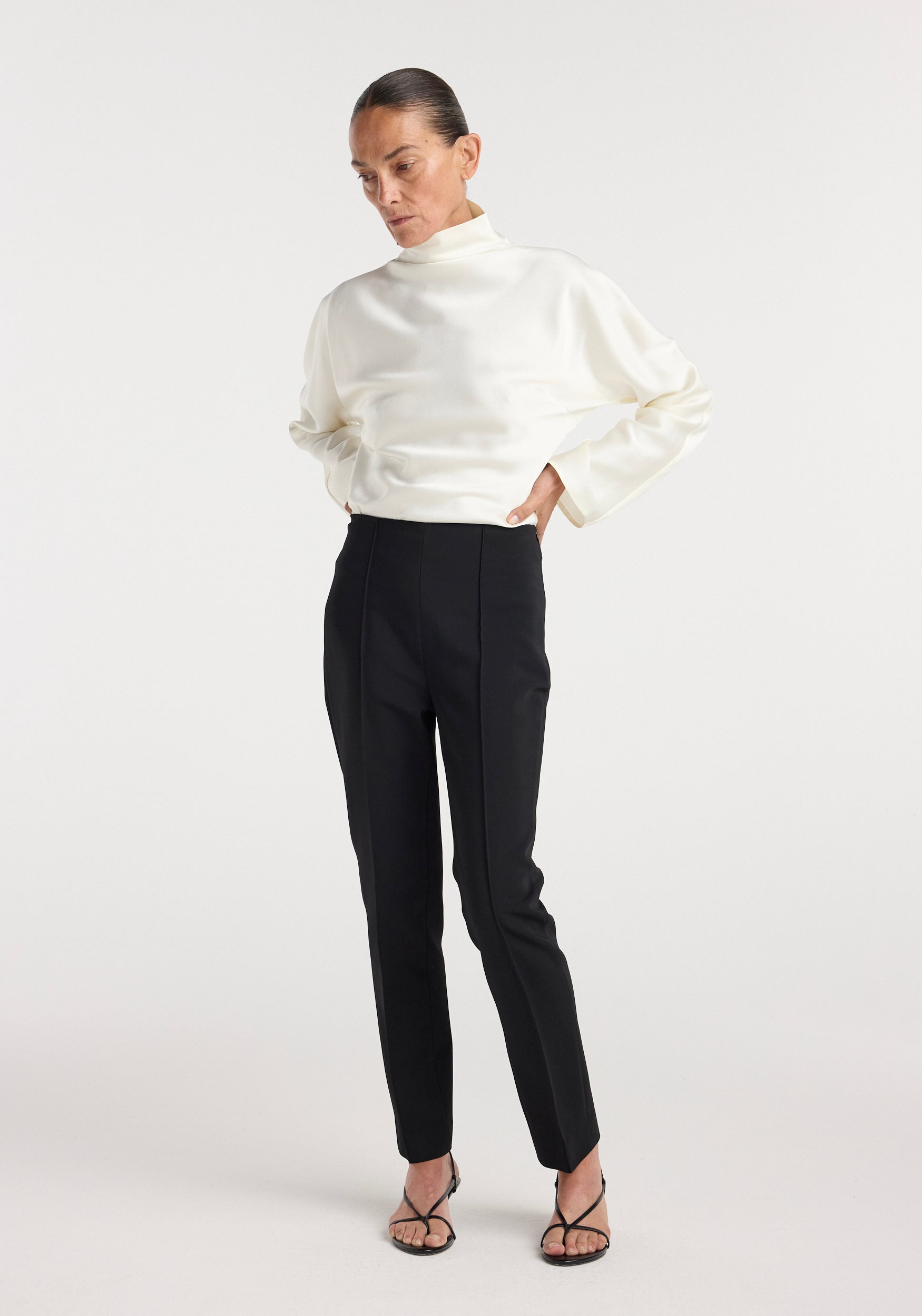 Woman wearing black Cigarette leg trousers | black paired with a white high-neck long sleeve top and black strappy sandals against a plain white background.