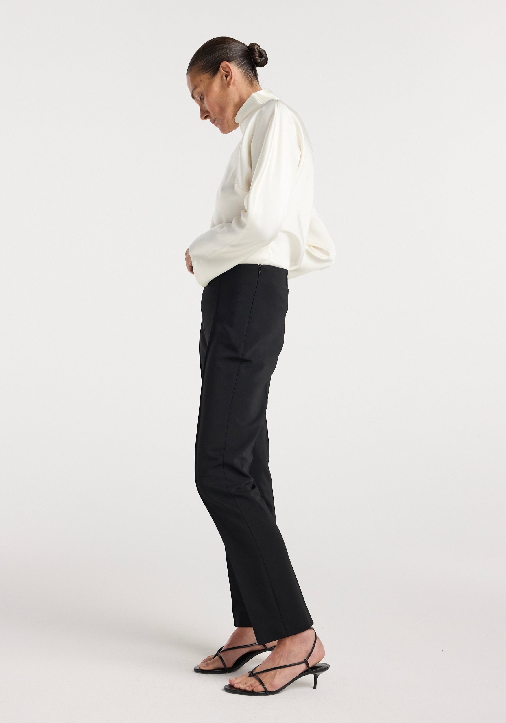 Side view of a woman wearing black Cigarette leg trousers | black paired with a white long-sleeve top and black strappy heels, standing against a plain background.