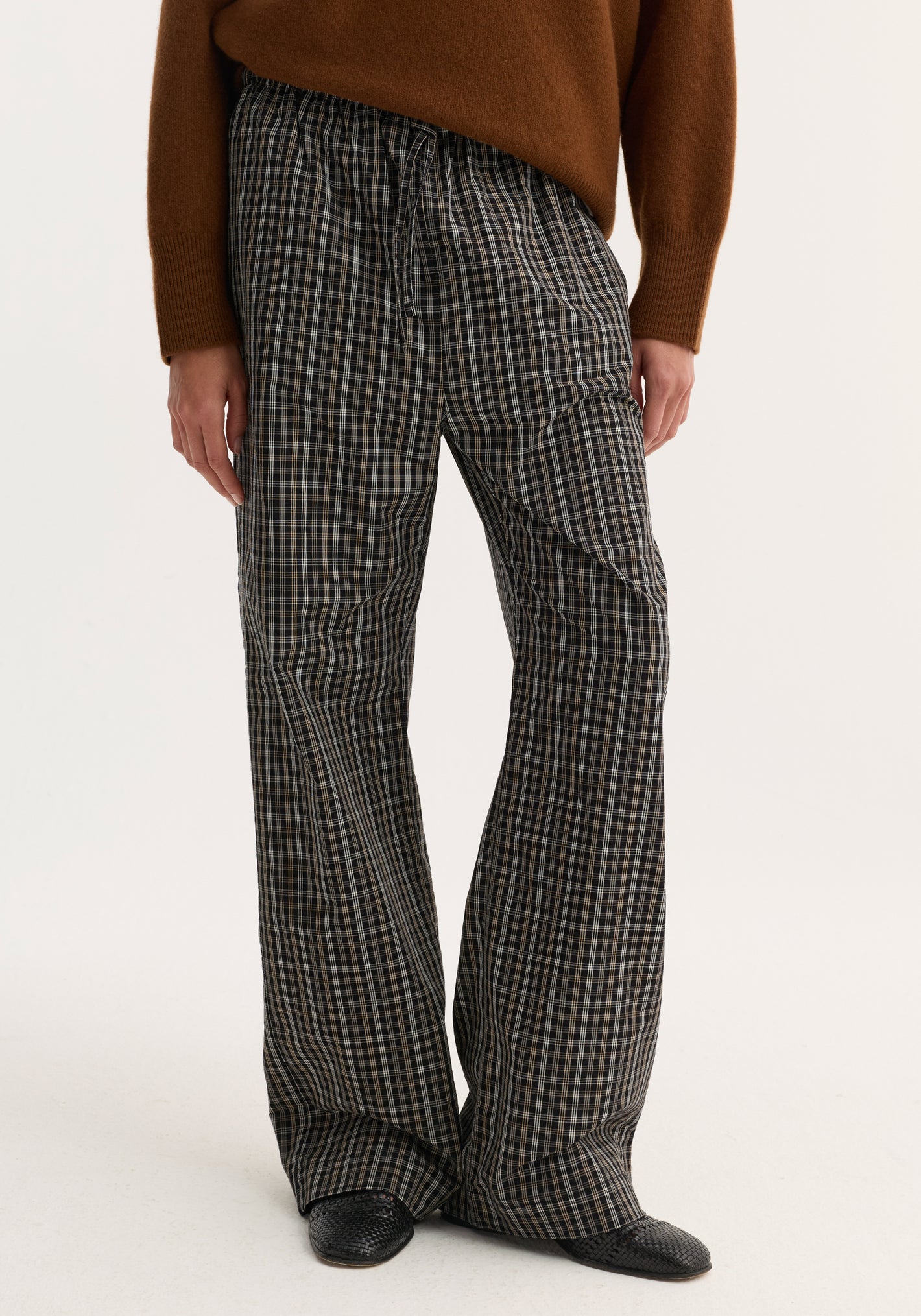 Relaxed fit trousers in black-and-white check, featuring a wide leg and drawstring waist. Styled with a brown sweater and black slip-on shoes.