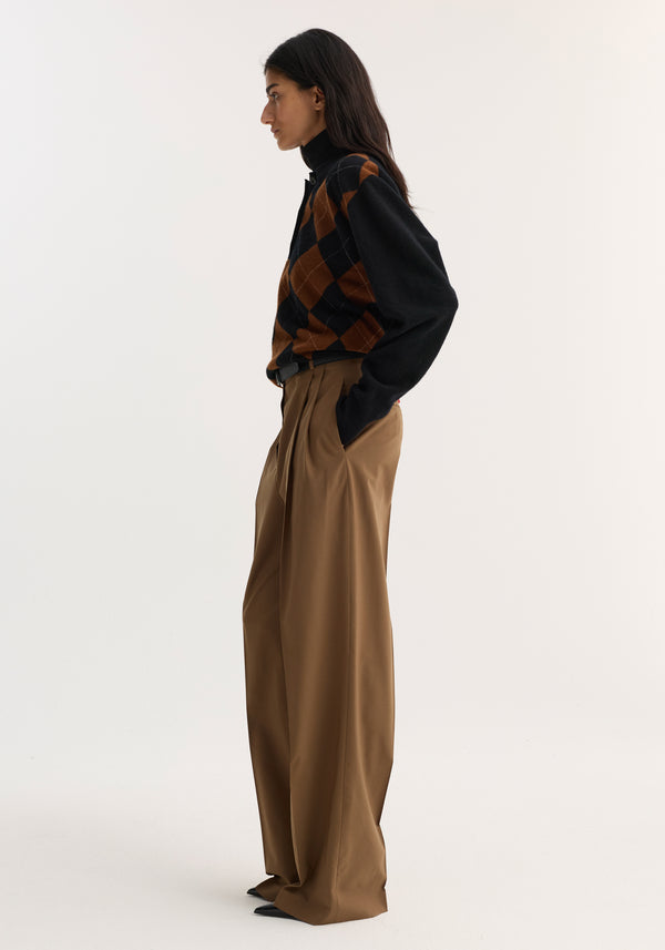 Person in profile wearing Mid-waist tailored trousers in mocha, showcasing a wide-leg, double-pleated design with a structured waistband, paired with a black turtleneck and argyle vest.