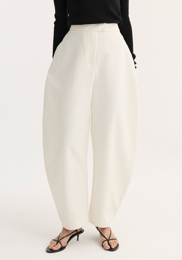 Model wears Sculptural trousers | paper white, high-waisted, wide-leg with tulip silhouette, ankle-length. Features discreet side pockets, styled with black top and sandals.
