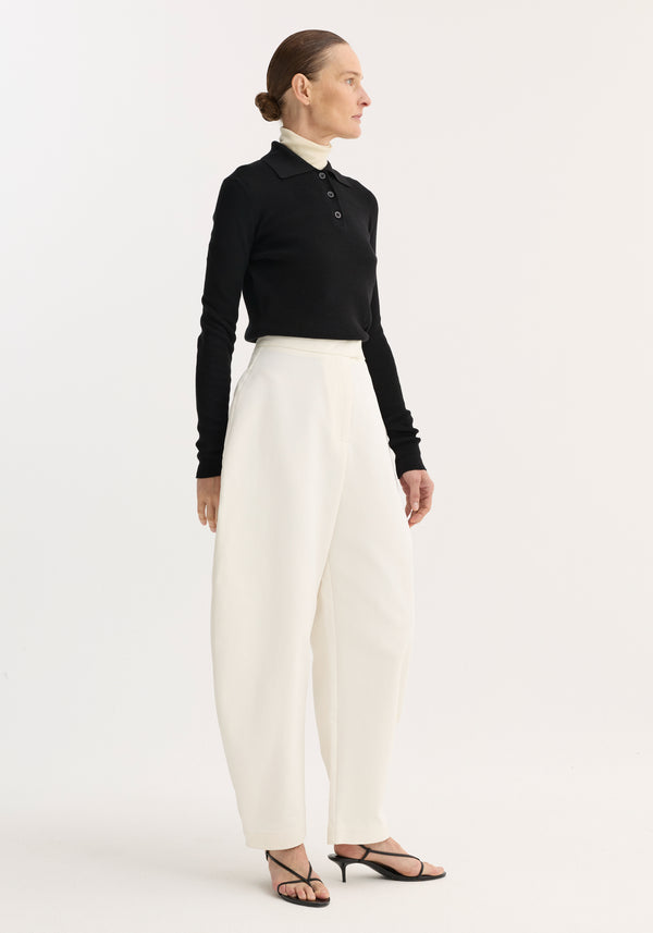Woman models sculptural trousers in paper white with a balloon leg and high waist, paired with a black rib-knit polo sweater over a white turtleneck.