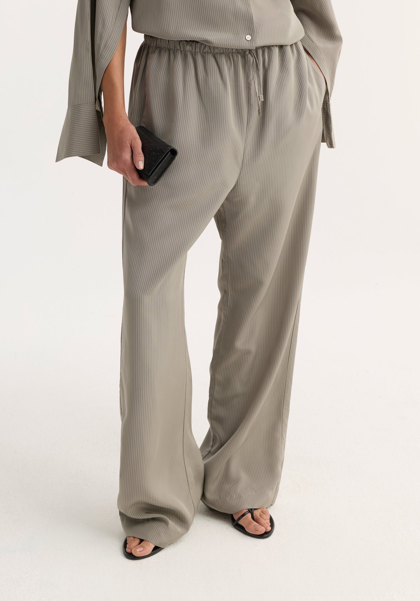 Relaxed fit trousers in light grey stripe, featuring a drawstring waist and wide leg, worn with matching top and black sandals, holding a black wallet.