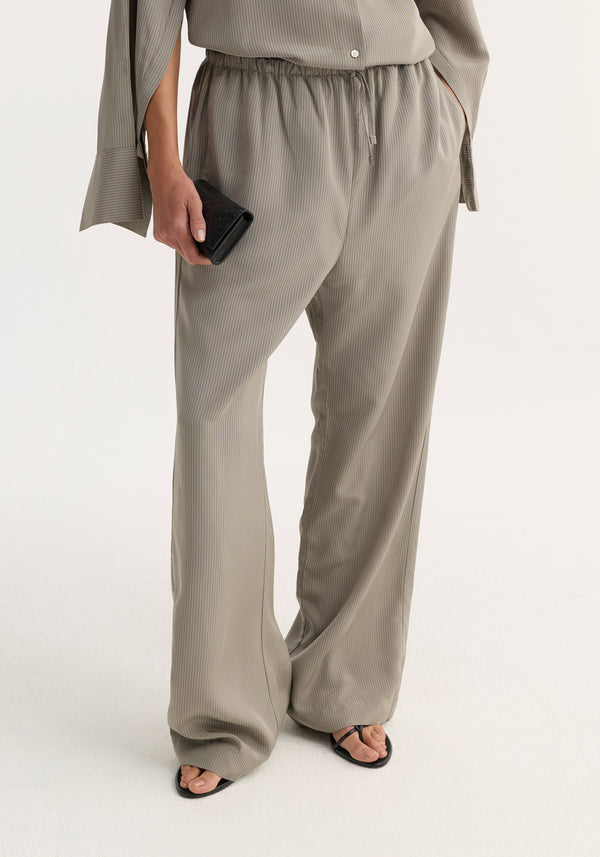 Relaxed fit trousers in light grey stripe, featuring a drawstring waist and wide leg, worn with matching top and black sandals, holding a black wallet.