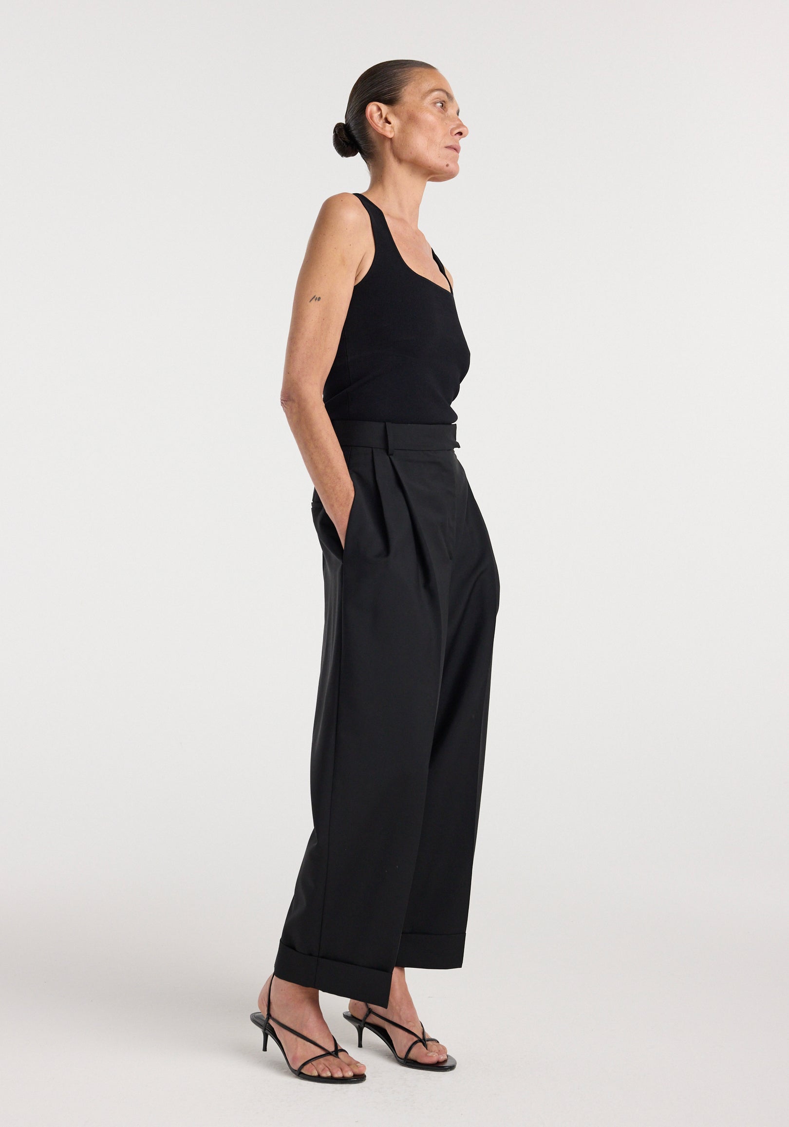 Woman modeling a sleeveless Bustier shaped knitted top | black, paired with high-waisted black cropped trousers and black strappy heels, standing in profile.