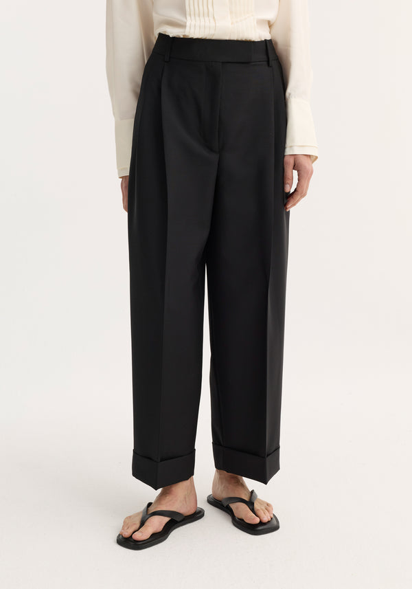 High-waist turn-up trousers in black, featuring sharp pleats and a wide leg, paired with black sandals. A cream blouse is tucked in.