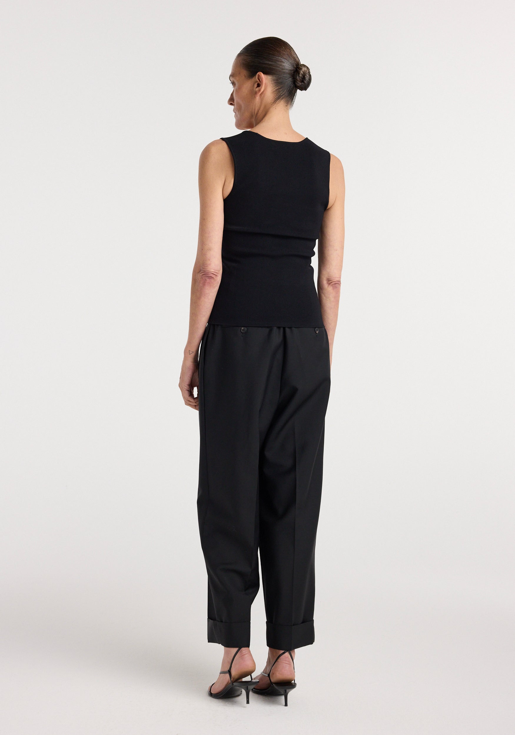 Woman seen from behind wearing a fitted sleeveless Bustier shaped knitted top | black paired with loose black trousers and strappy heeled sandals against a plain background.