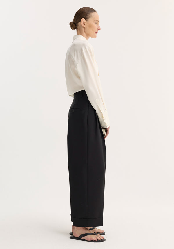 Side-profile of a model in High-waist turn-up trousers | black, showcasing wide-leg fit, pleating, and turned-up hem, paired with an ivory shirt.