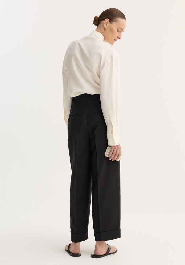 Person wearing high-waist turn-up trousers in black, showcasing wide legs and sharp pleats, paired with a relaxed ivory shirt and holding a cream clutch.