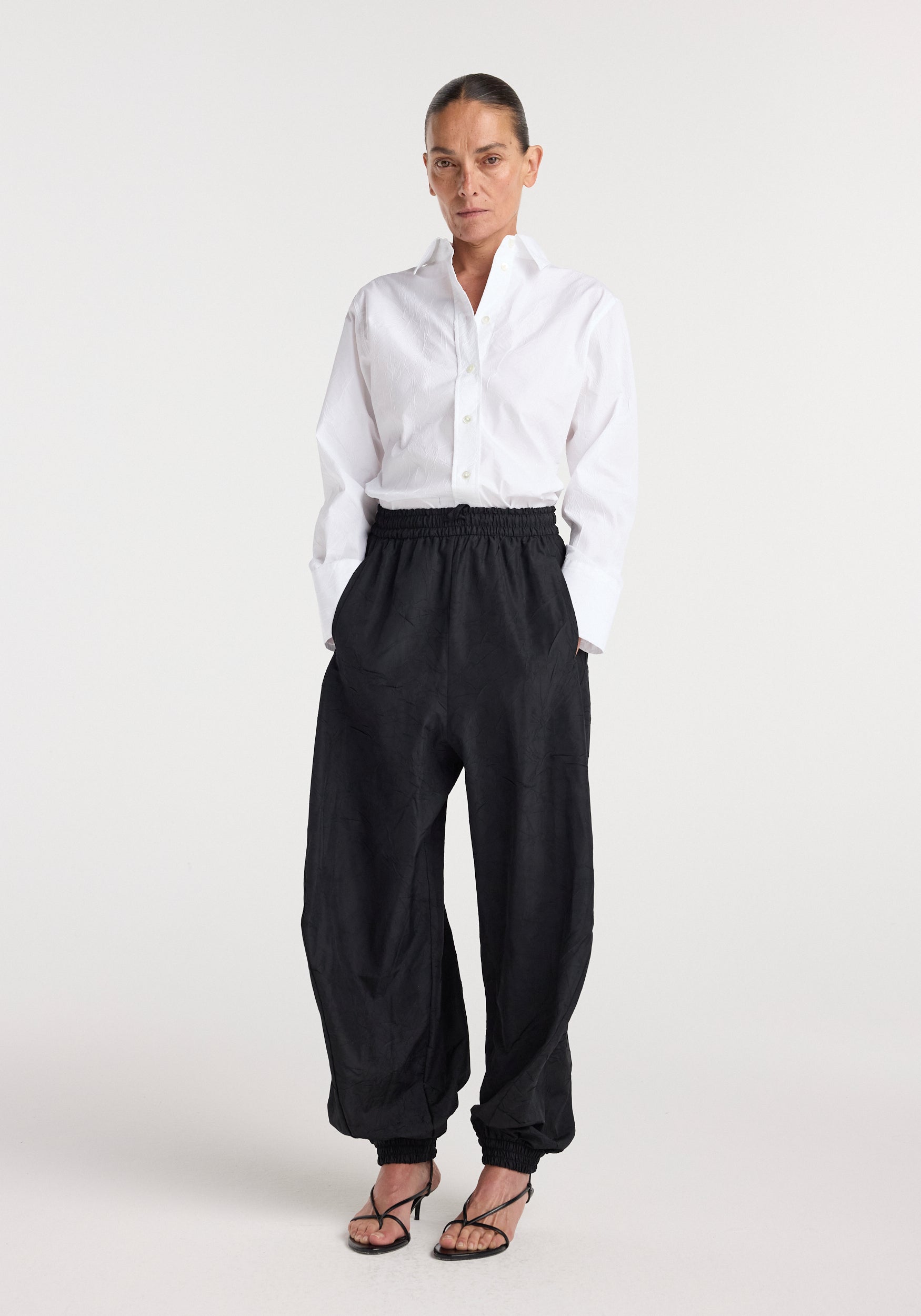 Woman wearing black Balloon track pants | black with an elastic waistband and cuffs, paired with a white button-up shirt and black strappy heels, standing against a plain white background.