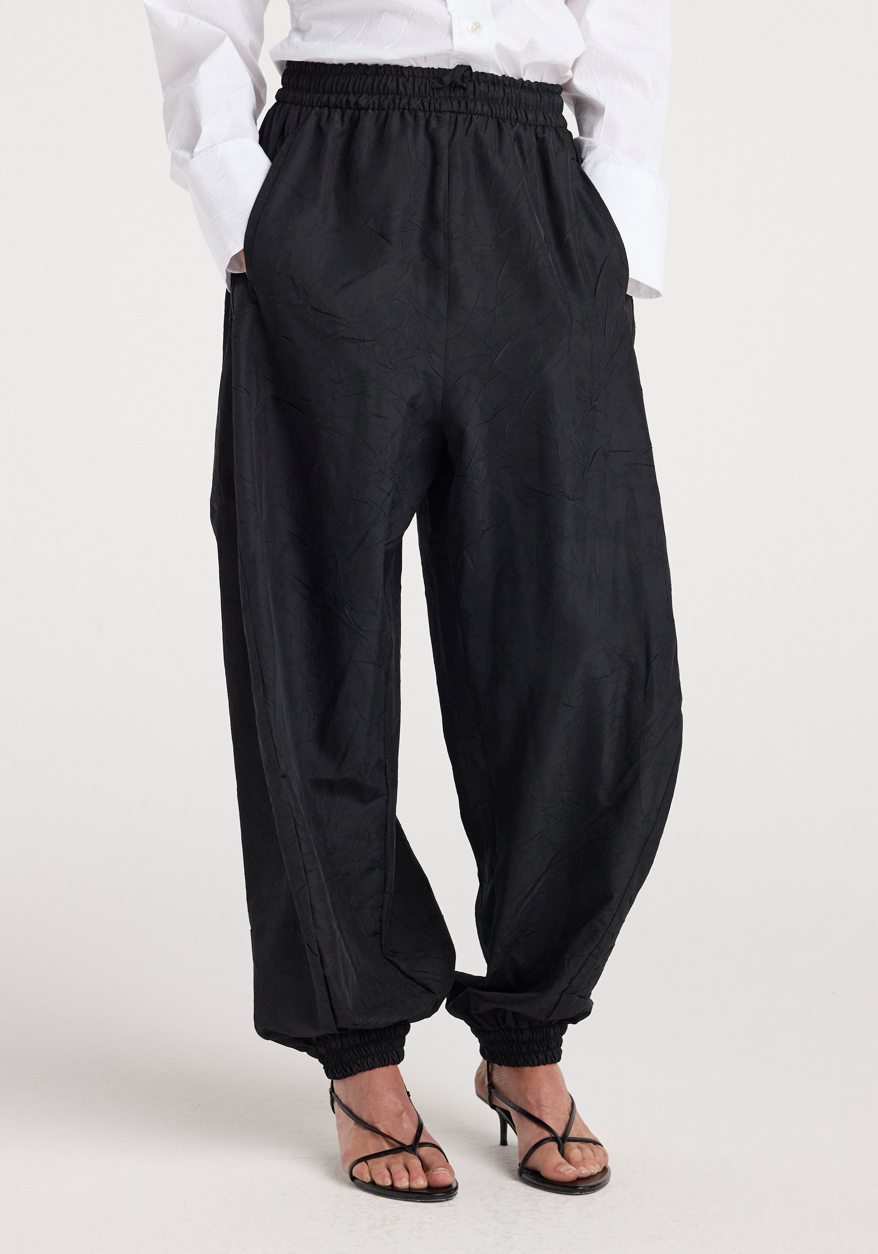 Front view of a person wearing black Balloon track pants | black with an elastic waistband and ankle cuffs, styled with a white shirt and black high-heeled sandals.