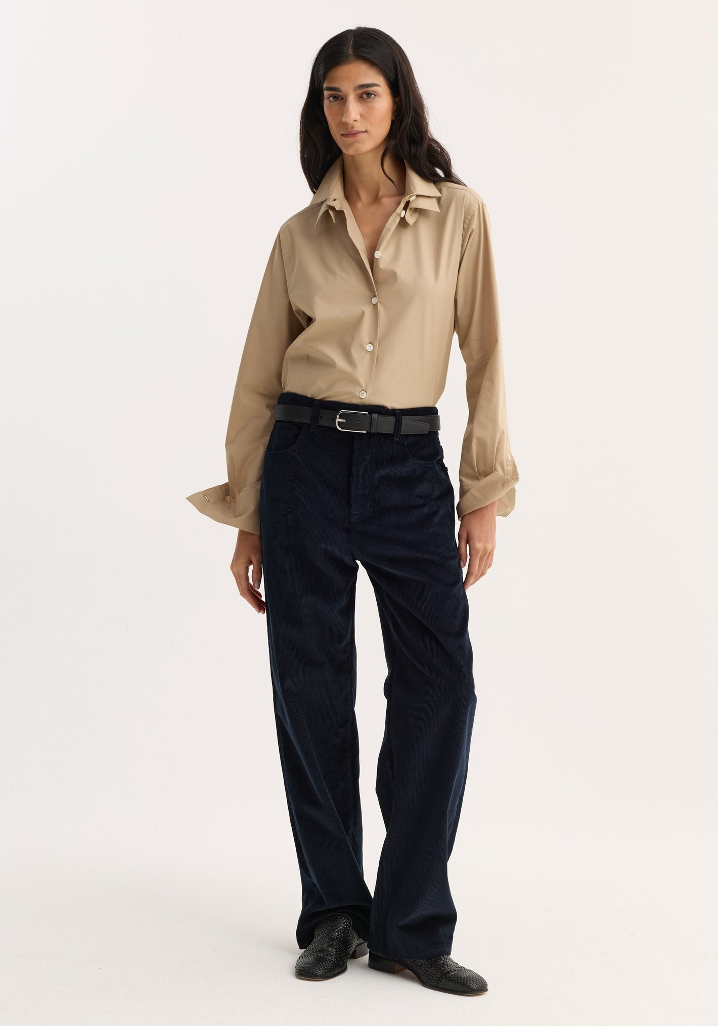 Woman wearing Relaxed corduroy trousers in navy, styled with a beige button-down shirt, black belt, and shoes, standing against a plain background.