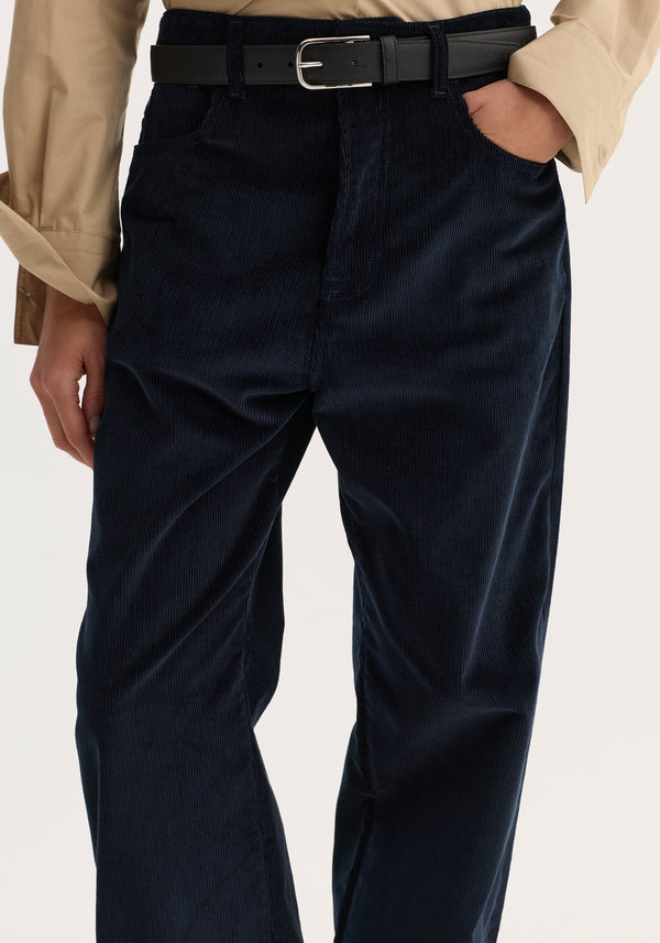 Relaxed corduroy trousers in navy, featuring a loose fit and denim-style details, worn with a black leather belt and beige long-sleeve shirt.