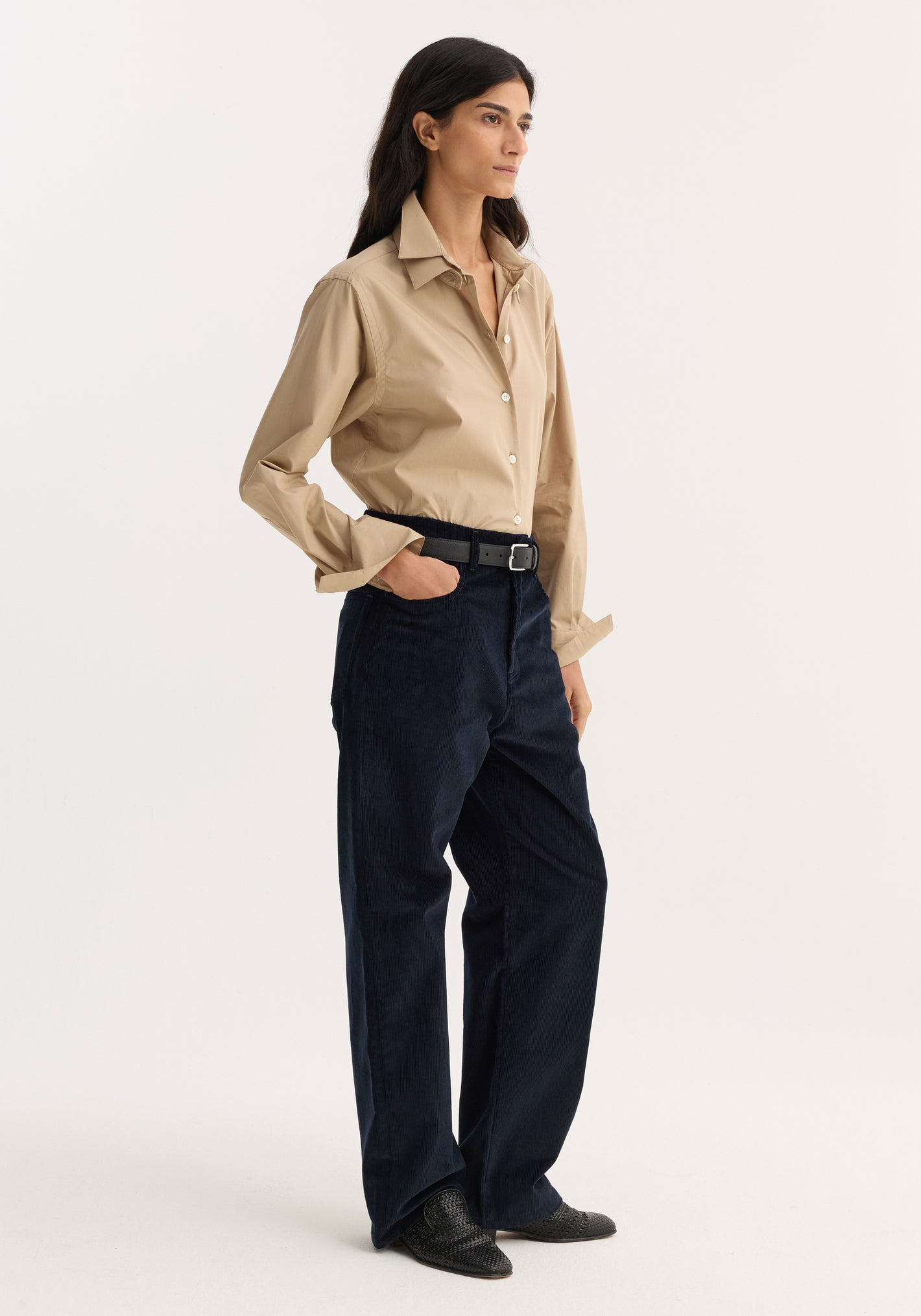 Woman wearing Relaxed corduroy trousers in night, styled with a beige shirt, black belt, and pointed-toe shoes, hand in pocket, looking right.