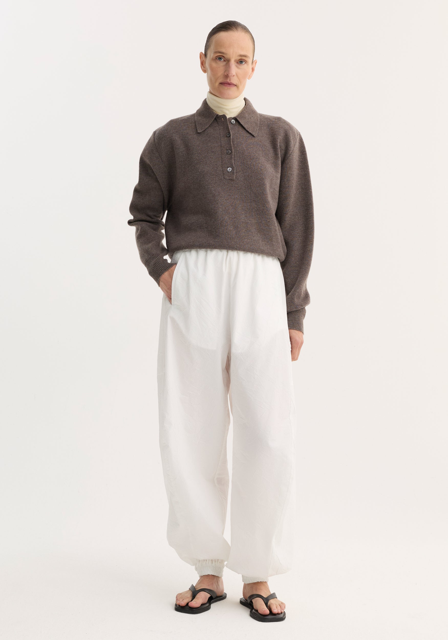 Model wearing Balloon track pants in optic white, featuring a voluminous leg, elasticated waistband, and front welt pockets, paired with a layered top and sandals.