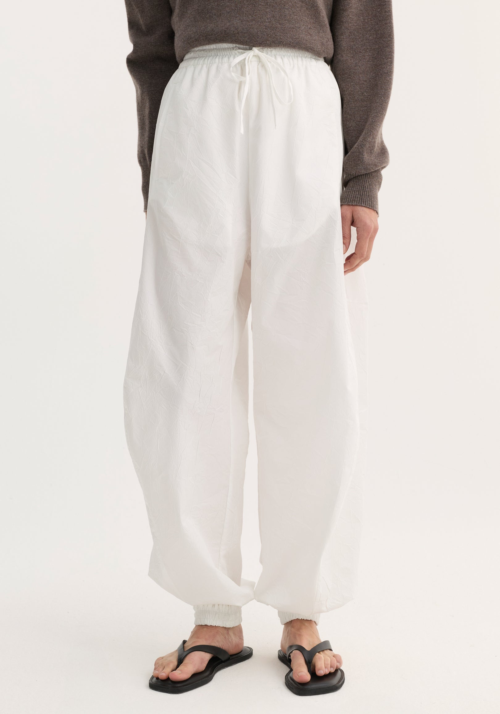 Balloon track pants in optic white with crinkled texture, elastic waistband, drawstring, and welt pockets. Modeled with black sandals, showing loose, voluminous leg design.