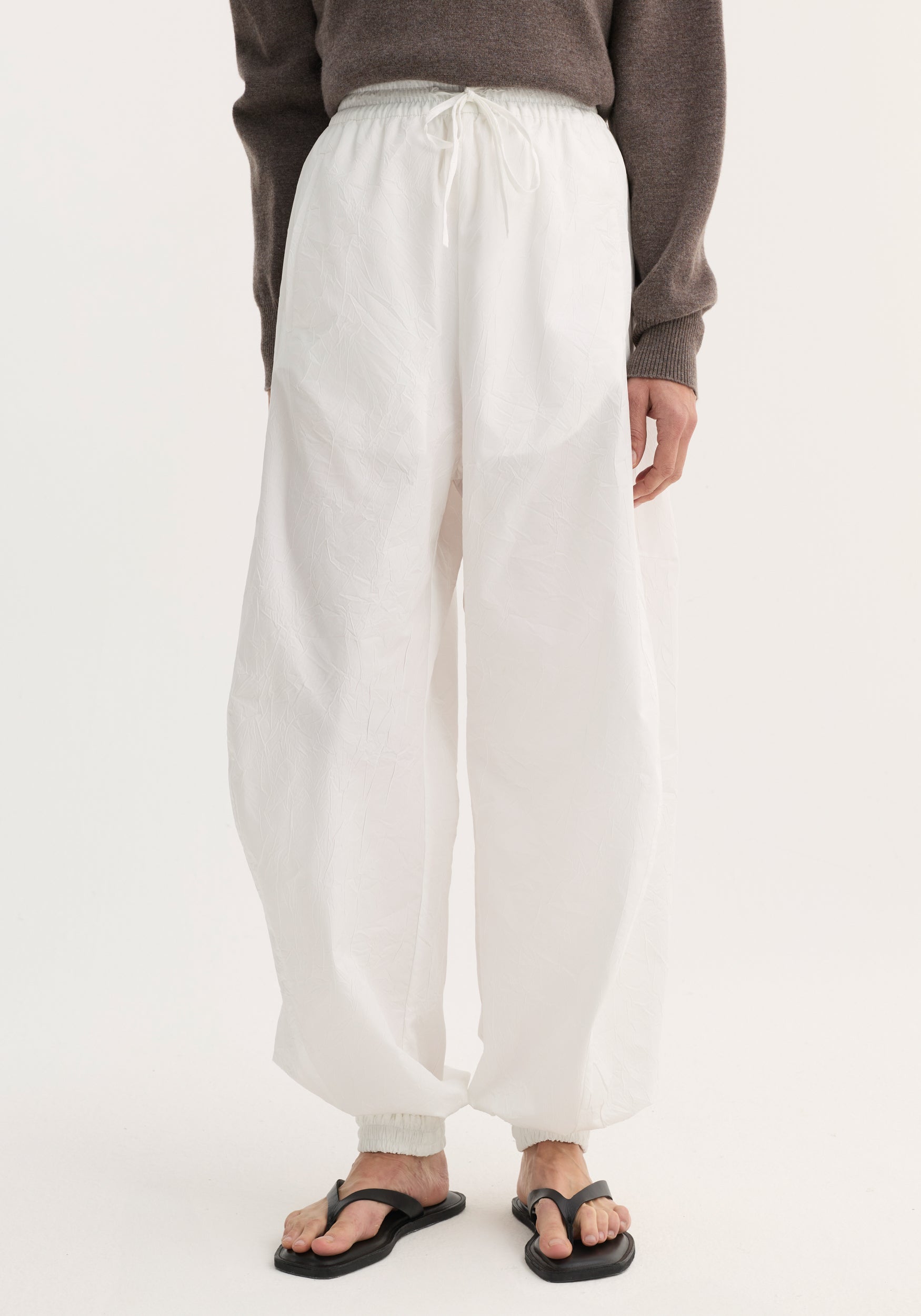 Balloon track pants in optic white with crinkled texture, elastic waistband, drawstring, and welt pockets. Modeled with black sandals, showing loose, voluminous leg design.