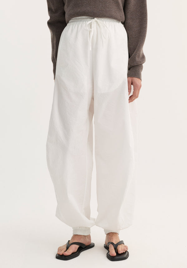 Balloon track pants in optic white with crinkled texture, elastic waistband, drawstring, and welt pockets. Modeled with black sandals, showing loose, voluminous leg design.