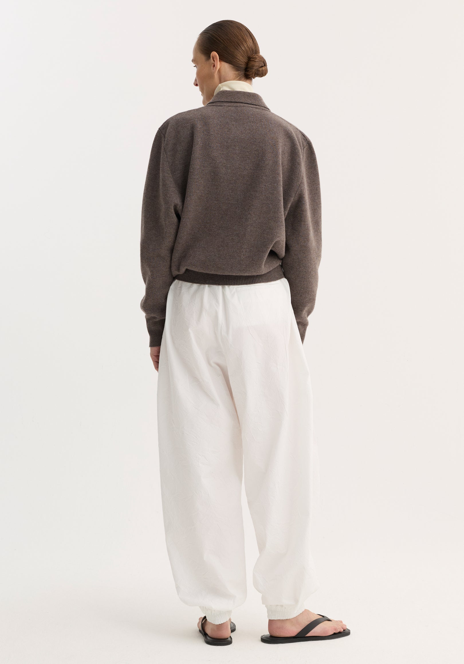 Person wearing Balloon track pants in optic white, showcasing their crinkled texture and voluminous leg, paired with a medium-brown sweater and black sandals.