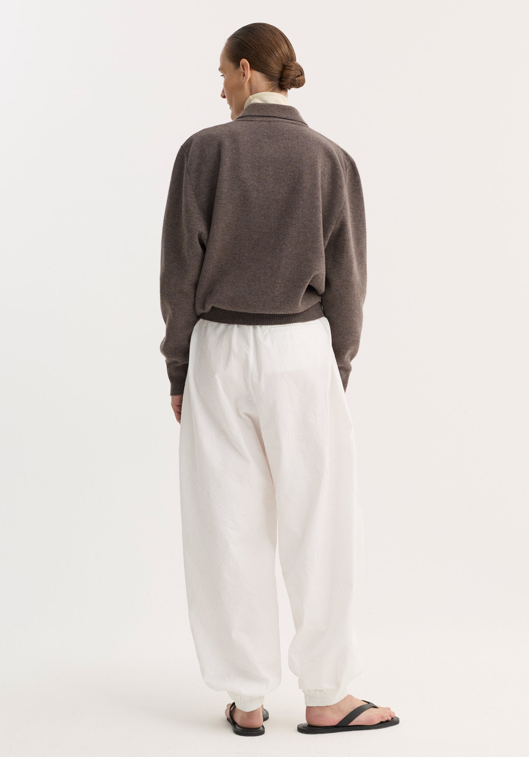 Person wearing Balloon track pants in optic white, showcasing their crinkled texture and voluminous leg, paired with a medium-brown sweater and black sandals.