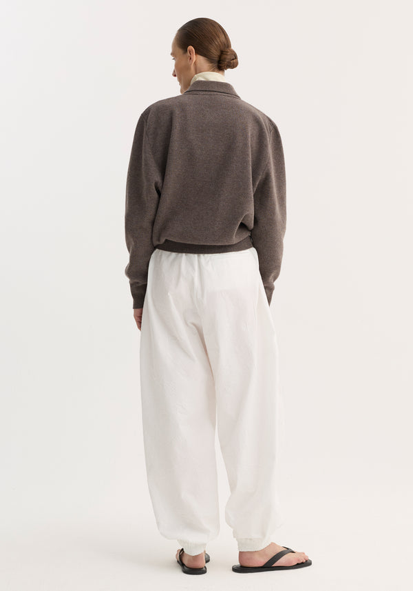 Person wearing Balloon track pants in optic white, showcasing their crinkled texture and voluminous leg, paired with a medium-brown sweater and black sandals.