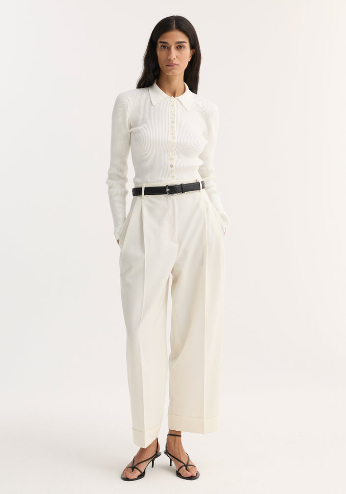Woman wearing high-waist turn-up trousers in off-white with sharp pleats, paired with a ribbed polo-style top, black belt, and strappy sandals.