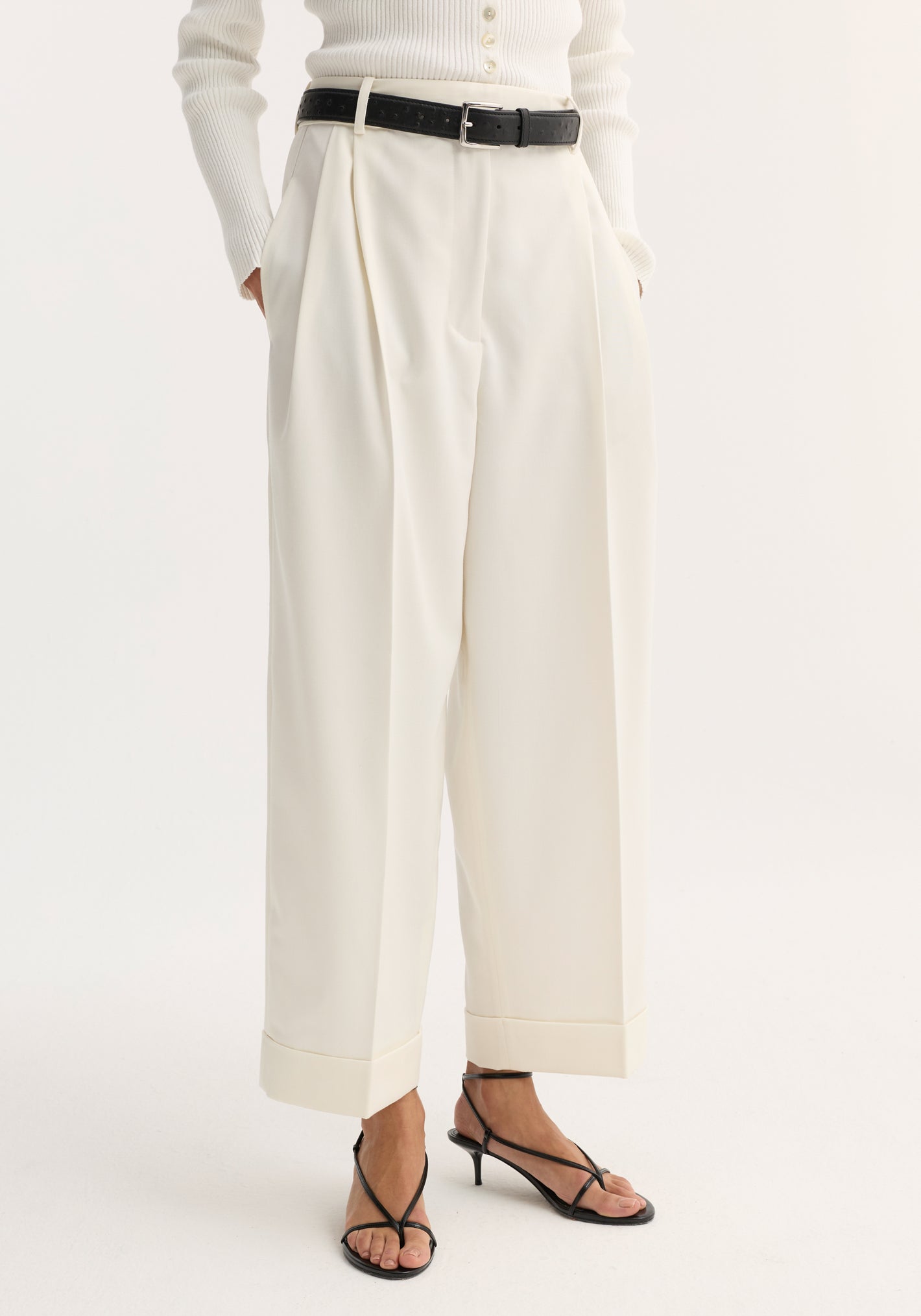 High-waist turn-up trousers in off white, featuring sharp pleats, wide legs, and a cropped, turned-up hem, paired with a black leather belt.