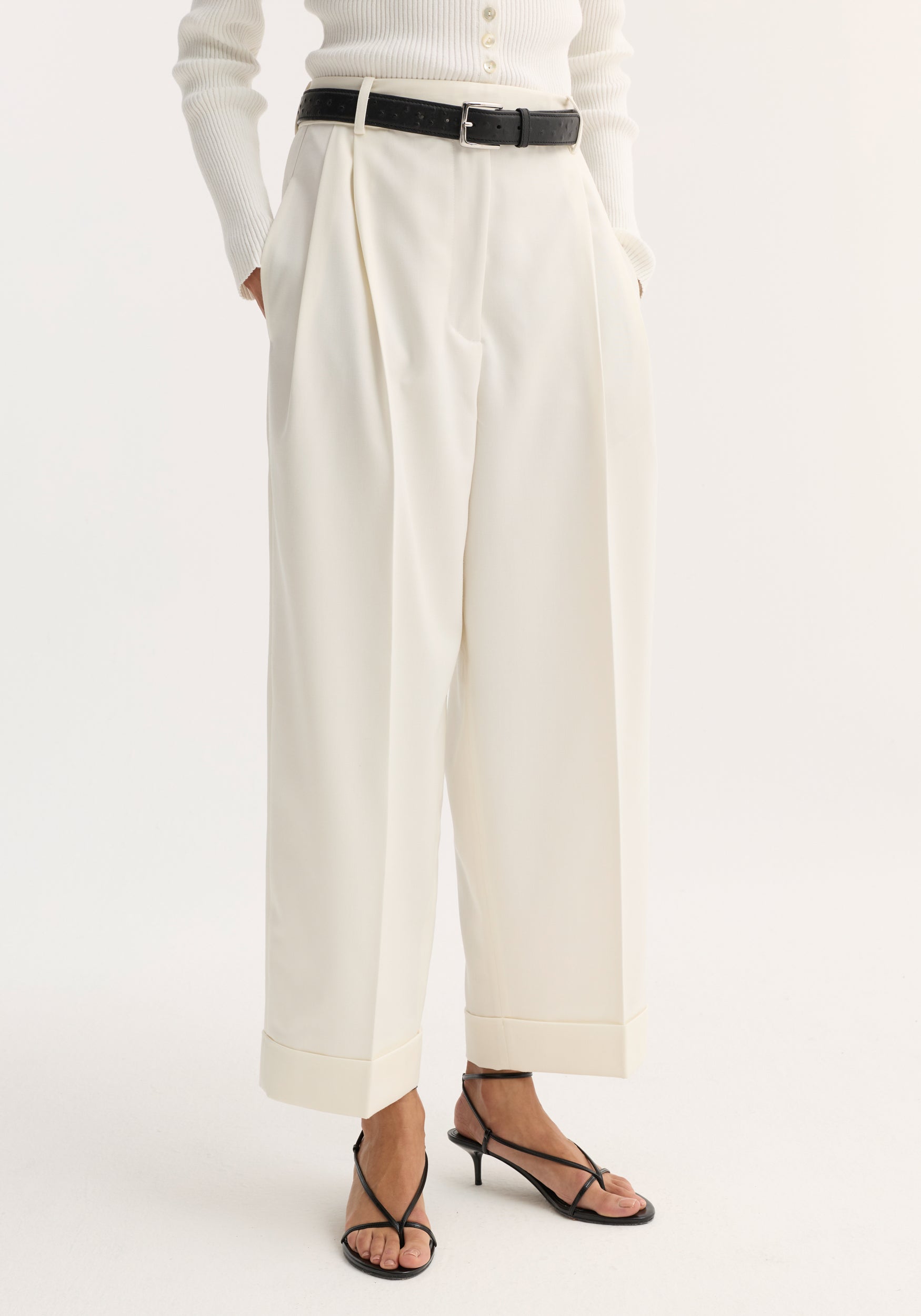 High-waist turn-up trousers in off white, featuring sharp pleats, wide legs, and a cropped, turned-up hem, paired with a black leather belt.