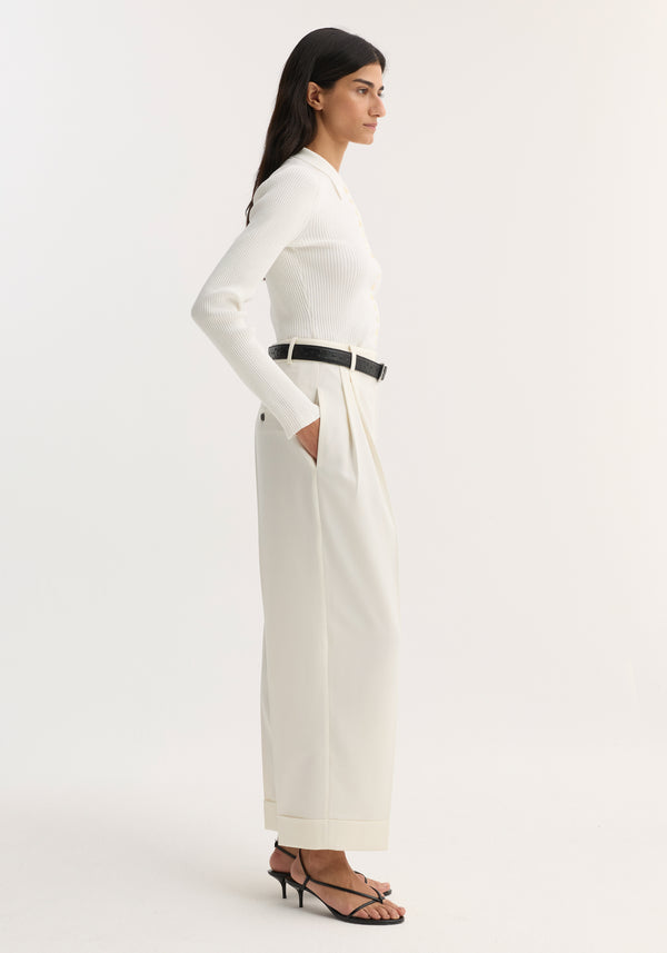 Model in high-waist turn-up trousers, off white, with pleats and side pockets, paired with a ribbed knit top, black belt, and sandals.