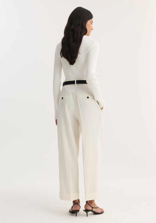 Model wearing high-waist turn-up trousers in off white, featuring sharp pleats and a wide leg, paired with a fitted ribbed top and black sandals.
