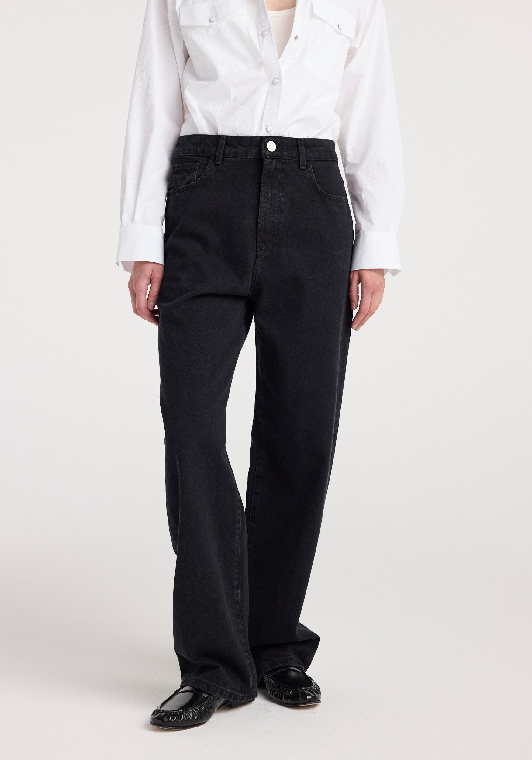 Front view of a person wearing black Bruun - Signature baggy denim | black paired with a white button-up shirt and black flat shoes, standing against a plain background.