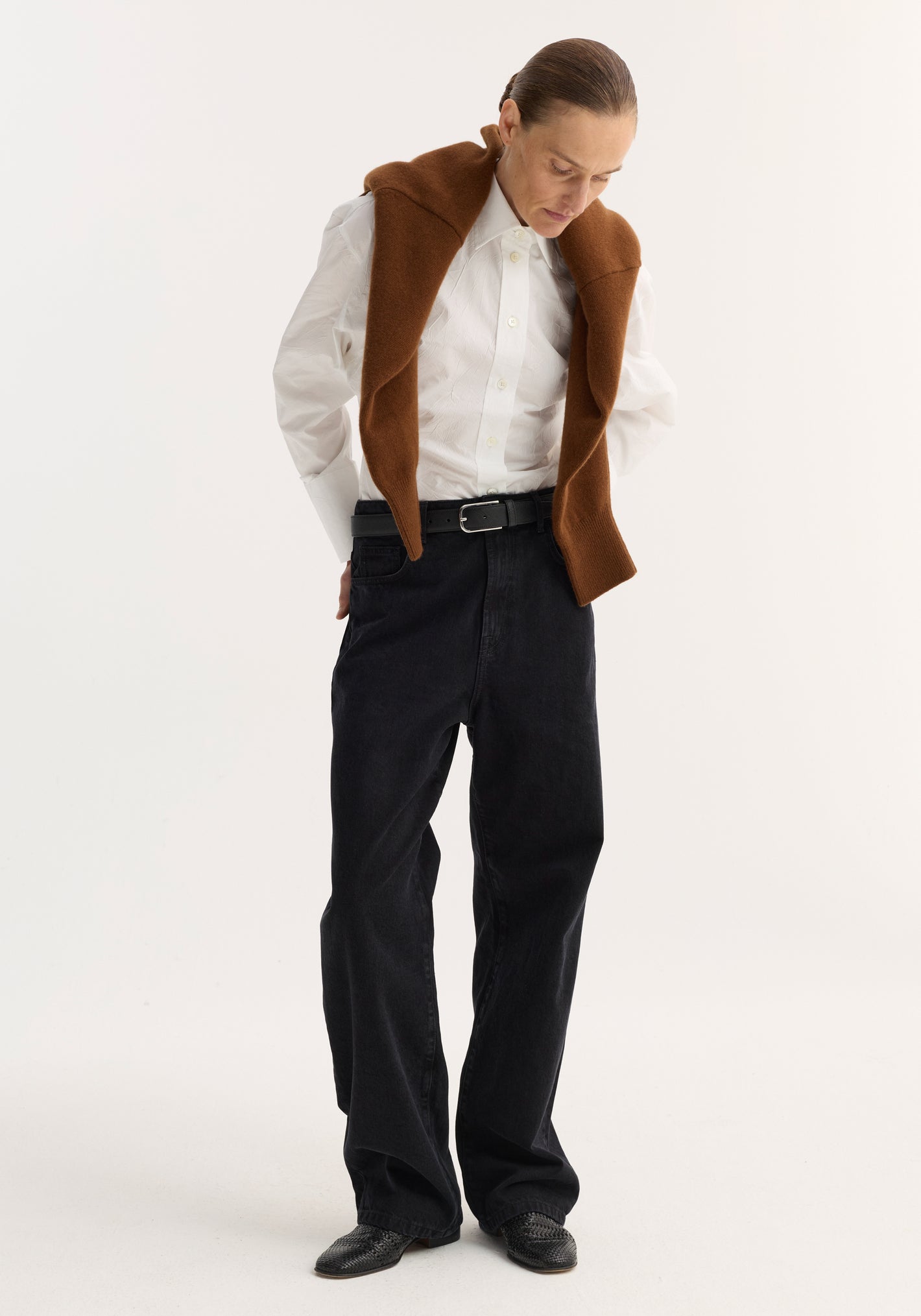 Person in baggy fit denim, black; low-waist, wide-leg jeans with a relaxed style, paired with a crisp button-down and loafers, sweater draped over shoulders.