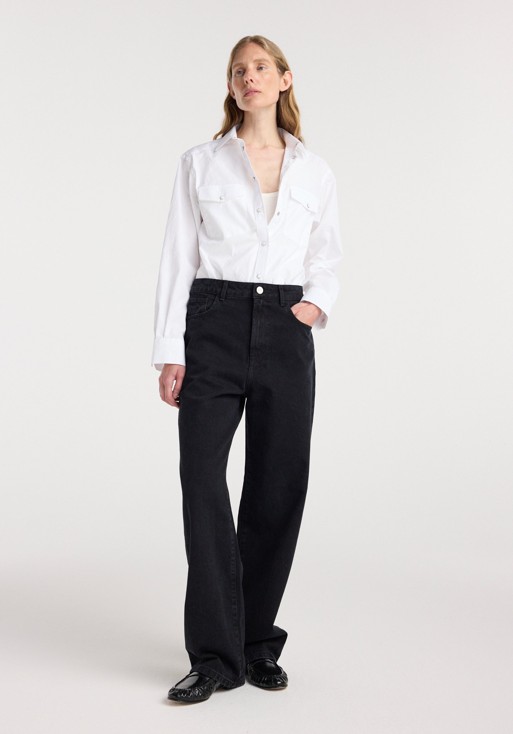 Woman wearing a white button-up shirt paired with Bruun - Signature baggy denim | black, standing with one hand in a pocket against a plain light background.