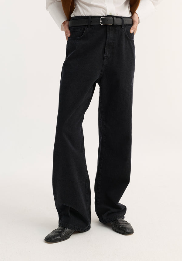 Person wearing Baggy fit denim | black with a high waist, black leather belt, black woven loafers, white shirt hem, and brown sweater draped over shoulders.
