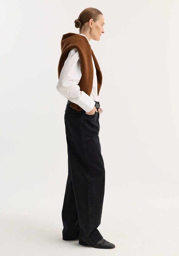 Person in profile wearing Baggy fit denim | black, showcasing low-rise, wide-leg design, paired with a white shirt, brown sweater, and black slip-on shoes.