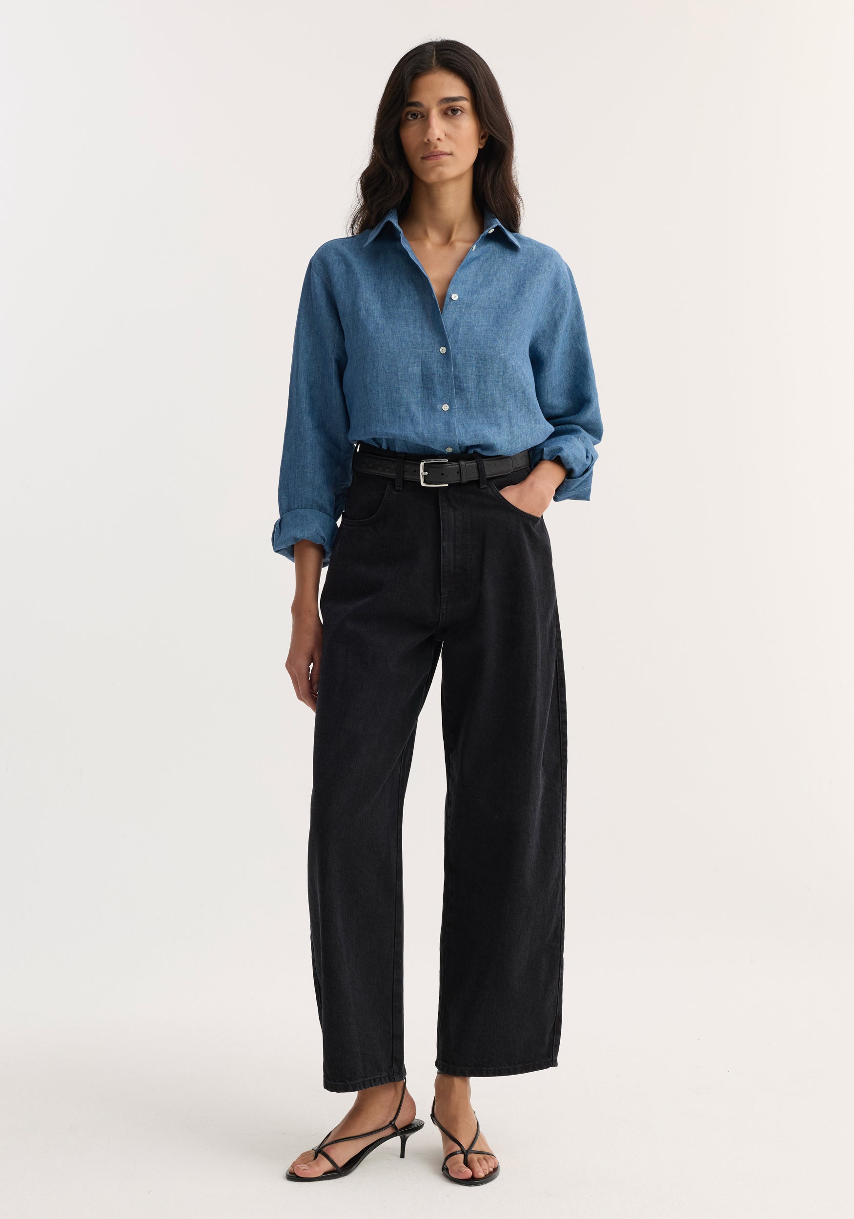 A woman wearing a blue denim shirt tucked into black Berber - Barrel leg denim | black jeans, paired with a black belt and black strappy sandals, standing against a plain background.