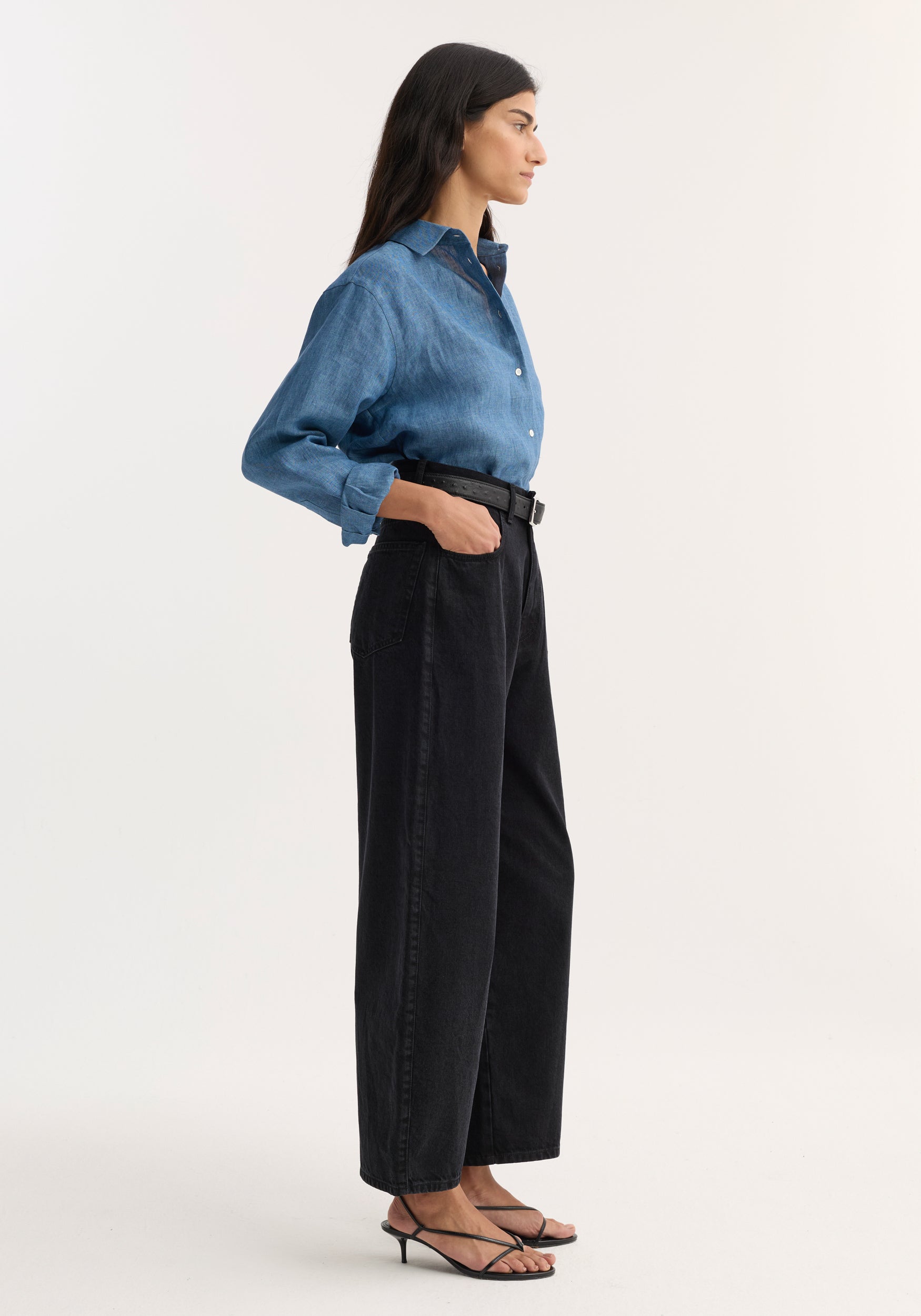 Profile view of a woman wearing Berber - Barrel leg denim | black with a tucked-in blue denim shirt and black belt, paired with black strappy heels against a plain background.