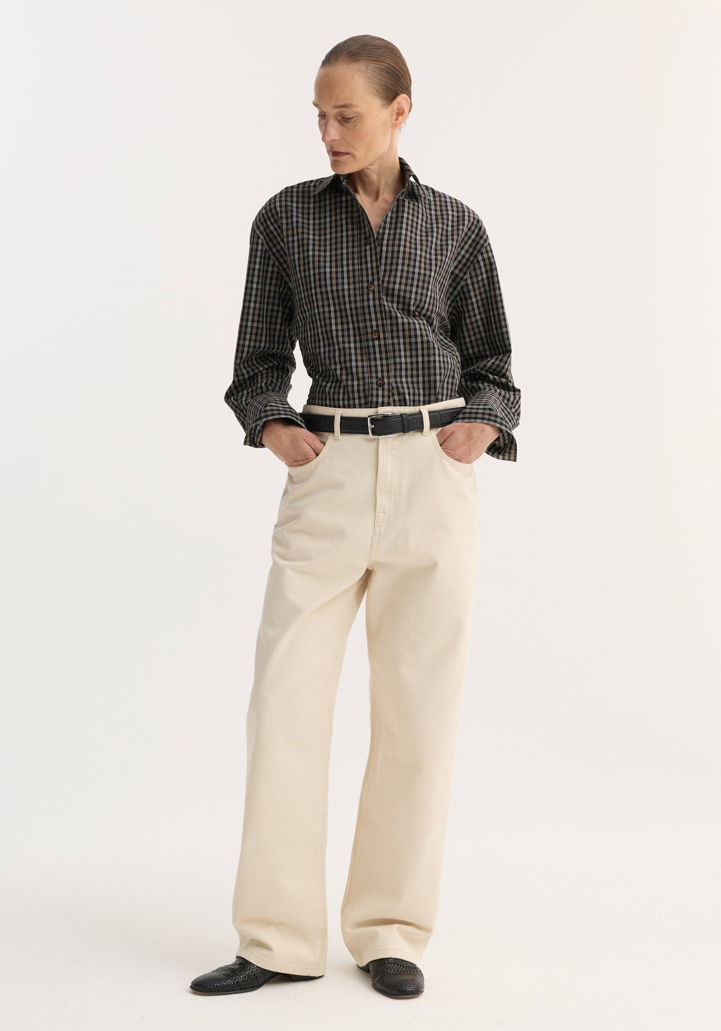 Person in plaid shirt and cream pants, showcasing Baggy fit denim | ecru with wide-leg design, hands in pockets, black shoes, minimal background.