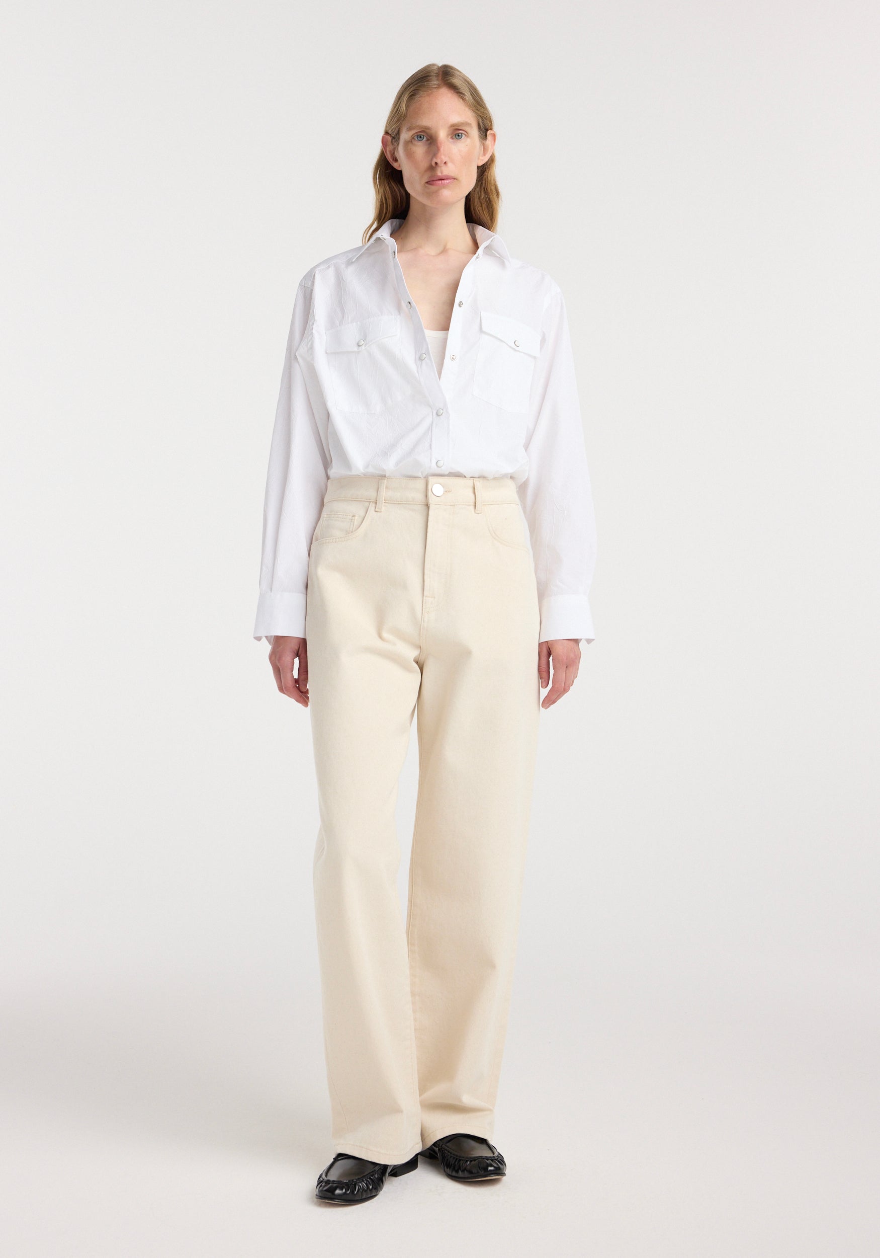 A woman wearing a loose white button-up shirt paired with Bruun - Signature baggy denim | ecru, standing against a plain light background with black shoes.