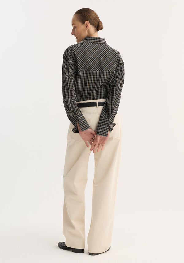 Person wearing Baggy fit denim | ecru, with wide-leg, high-waisted trousers, visible dark belt, and dark shoes. Hair in low bun, hands clasped behind.