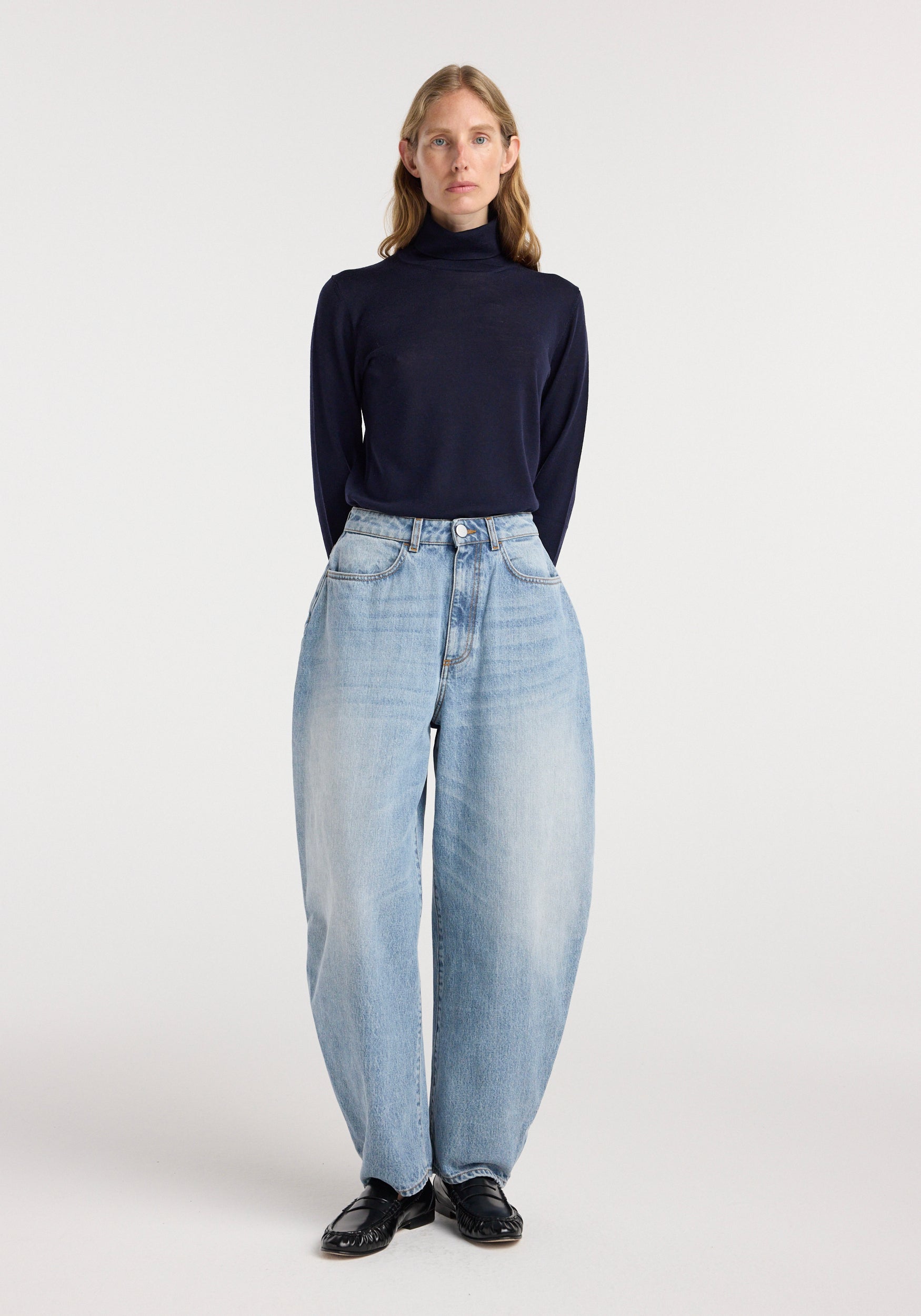 A woman wearing Fransje - Sculptural denim trousers | classic blue with a high waist and wide legs, paired with a fitted black turtleneck and black shoes, standing against a plain white background.