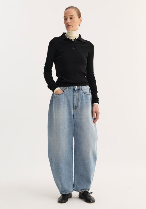 Person wearing sculptural denim trousers with a high waist and balloon legs, styled with a black polo top and black shoes. Jeans create a bold, modern silhouette.
